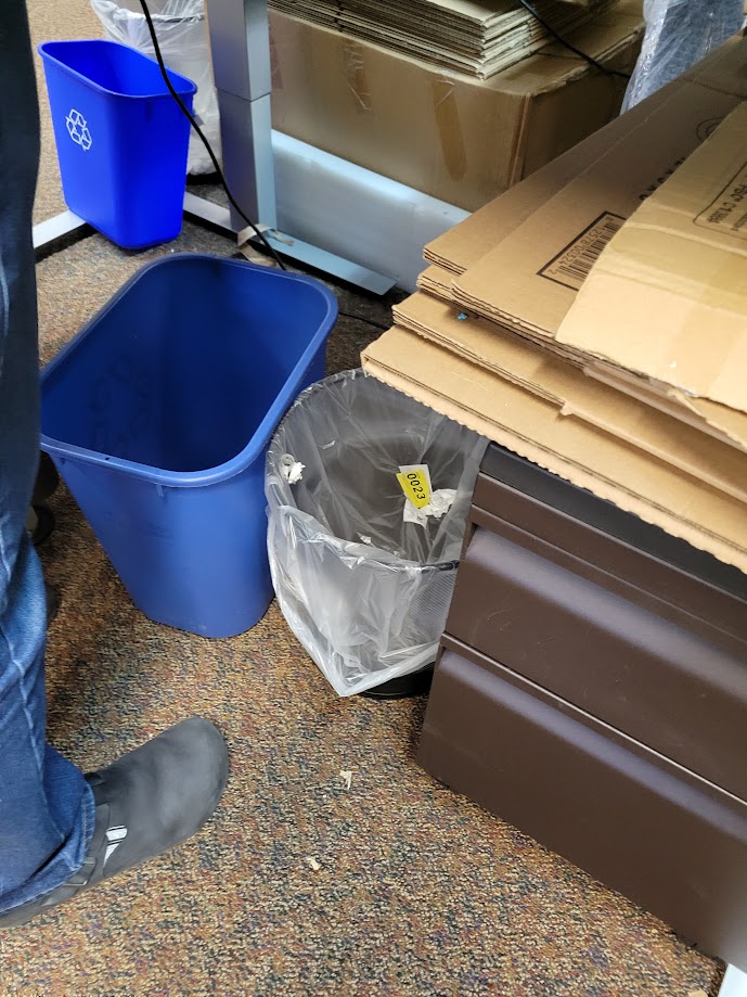 A wastebasket and recycle bin placed at the foot of a staff member's station.